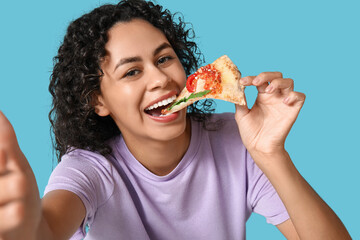 Beautiful young happy African-American woman eating slice of delicious pizza taking selfie on blue...