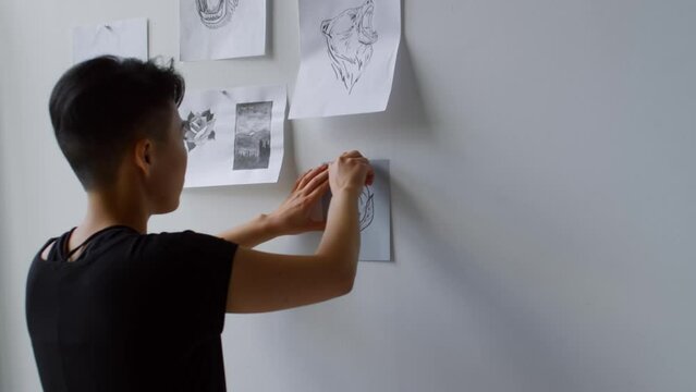 Young woman with short haircut hanging tattoo sketches on the wall with pins while working in studio