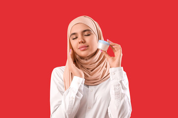 Young Muslim woman in hijab with jar of cream on red background