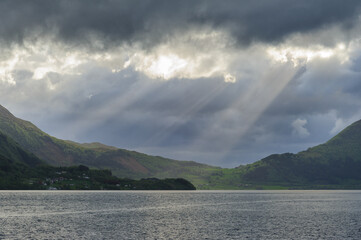 Sunbeams break through the dramatic cloud cover, illuminating parts of a serene lake surrounded by mountains as evening approaches.