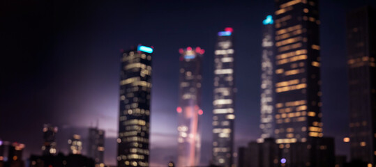 Fototapeta na wymiar Abstract night lights of a modern futuristic cityscape. Defocused image, ground level view of a dark modern urban city street with many tall buildings, towers, skyscrapers with glowing windows.