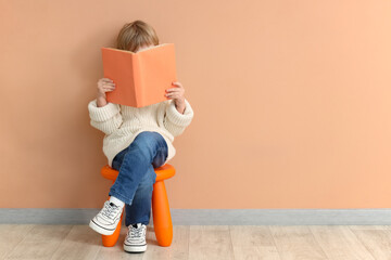 Cute little boy reading book while sitting on chair against color background