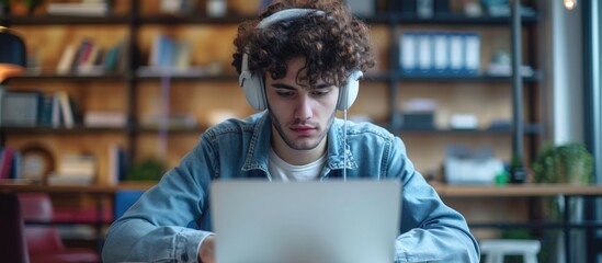 Student watching online lecture using laptop and headphones, taking notes in notebook. Curly-haired guy in office wearing earphones. Close-up. High-quality picture. - obrazy, fototapety, plakaty
