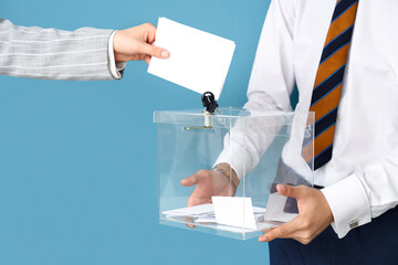 Hand putting voting paper into ballot box on blue background. Election concept