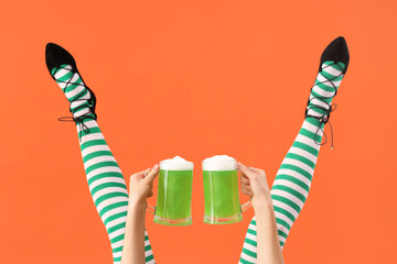 Female legs in green stockings with glasses of beer on orange background. St. Patrick's Day...