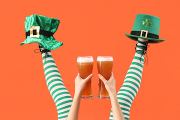 Female legs in green stockings with glasses of beer and leprechaun's hats on orange background. St....