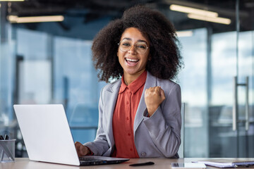 Portrait of a cheerful young businesswoman with curly hair enjoying success at her office workplace, showing excitement and confidence.