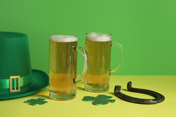 Mugs of beer with leprechaun hat, horseshoe and paper clovers on yellow table against green...