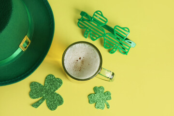 Mug of beer with leprechaun hat, plastic eyeglasses and paper clovers on yellow background. St....