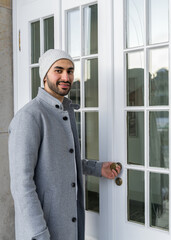 A smiling young man of Asian appearance in a light coat and a white knitted hat stands in front of the glass office door.