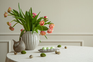 Minimalist composition of easter dining room interior with round table, vase with tulips, bowl with colorful eggs, hen sculpture, beige wall with stucco and personal accessories. Home decor. Template.
