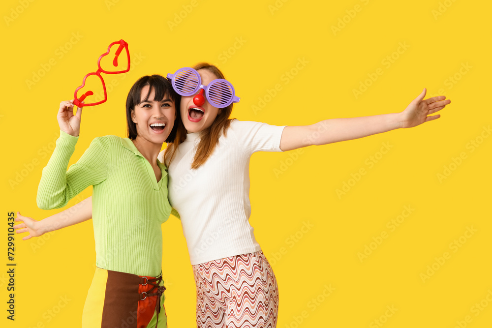 Wall mural beautiful young happy women in funny glasses on yellow background. april fools day celebration