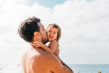 Cheerful young father hugging and kissing little kid against cloudy sky on sunny day