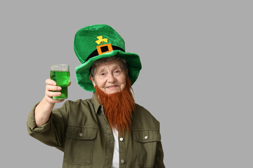 Senior woman in leprechaun hat with red beard holding glass of beer on grey background. St....