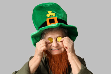 Senior woman in leprechaun hat with red beard holding coins on grey background. St. Patrick's Day celebration