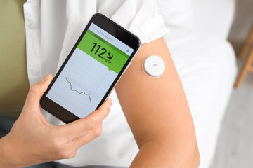 Diabetic woman with glucose sensor using mobile phone for measuring blood sugar level in bedroom, closeup
