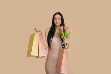 Beautiful young woman with shopping bags and bouquet of tulips on beige background. International Women's Day