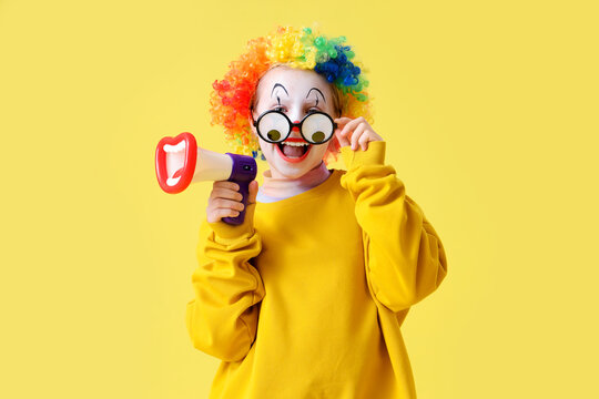 Funny little girl in clown wig with eyeglasses and megaphone on yellow background. April Fools' Day celebration