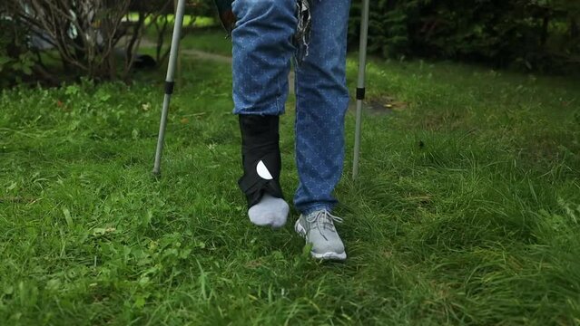 Woman with crutches. Crutches. Woman wearing a leg brace with adjustable side panels to immobilize and support her leg after surgery walking on crutches outdoors on a walkway in a garden. High quality