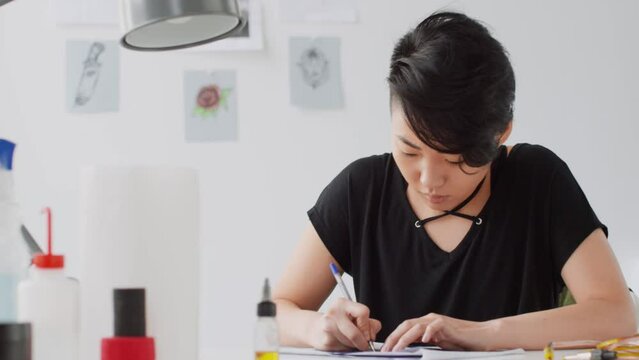 Young female artist sitting at desk in tattoo studio and drawing sketch with pen