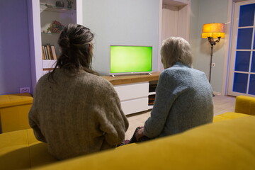 Family watching TV. Green screen. Grandmother and adult grandson are sitting on the couch. In front of them is a green screen TV. 

