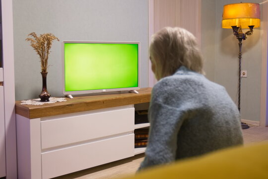 Grandma is watching TV. Green screen. Grandma is sitting on the sofa at home. In front of her is a green screen TV.