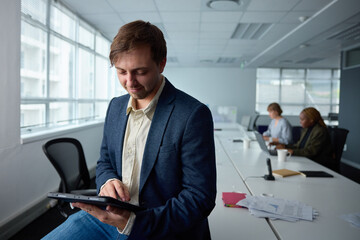Young businessman in businesswear sitting on desk while using digital tablet in office