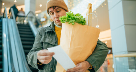 Price rise concept. Surprised male customer holding paper bag with food products looks at paper...