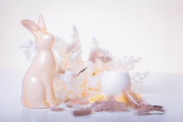 White Easter bunny next to an egg on a light background surrounded by boho-style feathers.