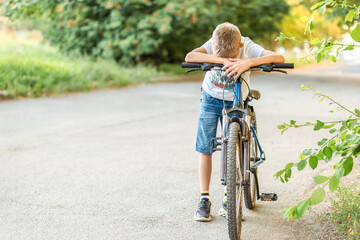 Naklejka premium Tired boy in a t-shirt and denim shorts leans on his bicycle after a long bike ride in the park, showing exhaustion and satisfaction