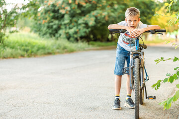 Naklejka premium Tired boy in a t-shirt and denim shorts leans on his bicycle after a long bike ride in the park, showing exhaustion and satisfaction
