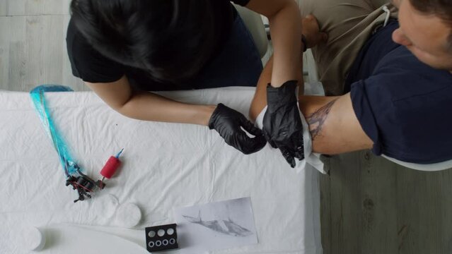 Top view of female tattoo artist wiping off excess ink from skin of male client with paper towel after making whale tattoo on bicep