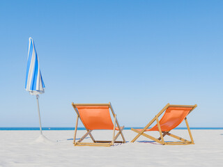 tranquil beach with two orange beach chairs and a blue-striped umbrella on a pristine sandy shore, with a clear blue sky and horizon in the background. Serene vacation concept.