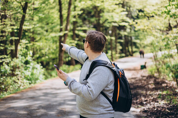Happy men with phone walking on city park and checks smartphone. Blonde male in sport clothes with backpack pointing finger outdoors, he hiking on woodland
