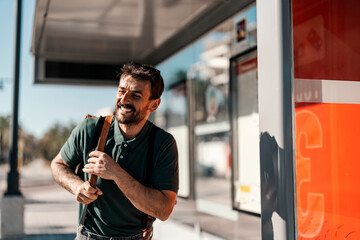 A happy young man is standing at bus stop and waiting for a bus.