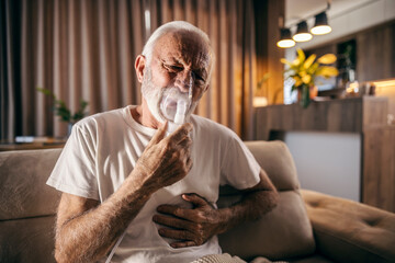 A sick senior man with pain in his lungs sitting at home and inhaling therapy.