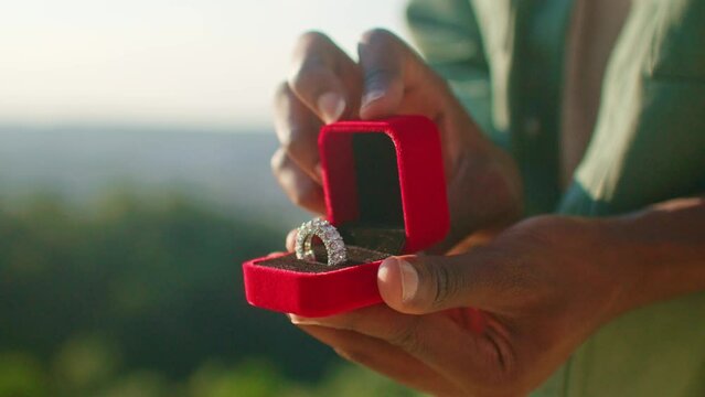 Very close camera view of African American male opening red box with large ring inside. Very big ring with many diamonds. Concept of proposal. Expensive jewelry. Wonderful landscape in background.