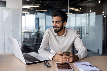 Smiling young Indian male businessman working in the office, sitting at a desk, holding a mobile...