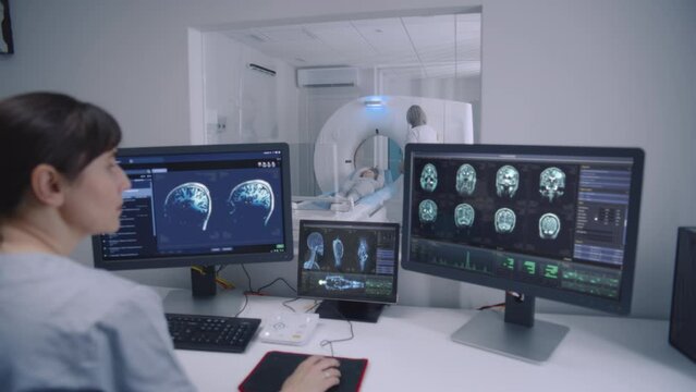 Female radiologist sits in control room and watches at monitors with displayed brain scans results. Professional doctor controls MRI or CT or PET scan with patient undergoing procedure at background.