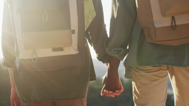 Rear view of mixed-raced couple wearing touristic backpacks while travelling. Couple in love holding hands and considering beautiful landscape or sunset. Happy together. Trust and love concept.