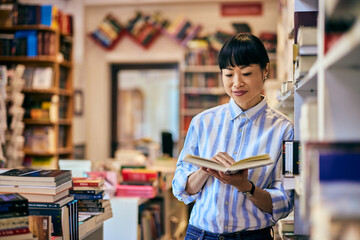 Asian woman standing, leaning on the bookshelf while reading one book in the library.