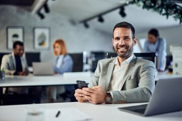 A portrait of a smiling businessman looking at the camera while using a phone and sitting in the co-working space.