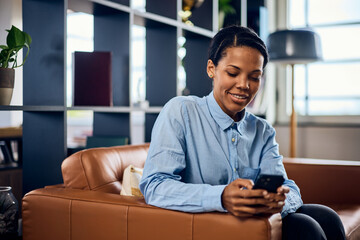 A smiling African woman, sitting on the sofa in her modern office and using a mobile phone.
