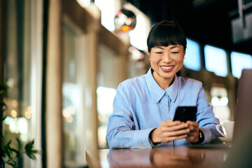 A black-haired woman using a mobile phone, sitting at the cafe, smiling.