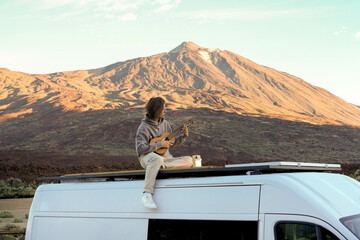 A young man sits on top of a van roof, playing a guitar against the scenic backdrop of Teide...