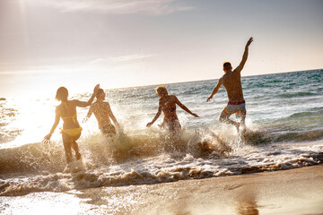 Four happy young friends or tourists are having fun, runs and jumps at sunset sea beach. Tropical...