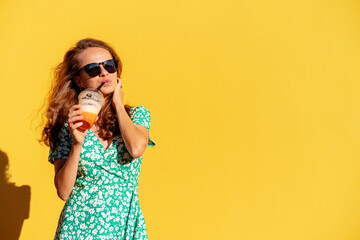 Happy young woman in summer green dress in sunglasses drinks juice and posing in front of yellow...