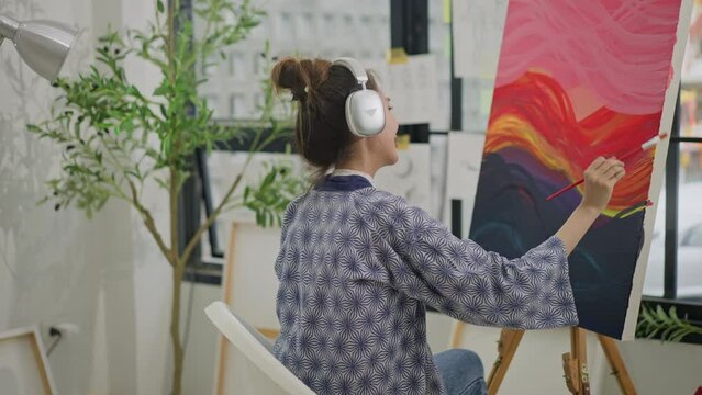Asian woman artist listen music with headphones while painting picture on canvas creating artwork,holding tube of oil paint,fine art artist drawing at art studio.asian female painting brush on canvas
