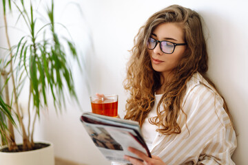 Young woman sitting on floor, drinking tea and reading book. Lifestyle, relaxation and domestic...
