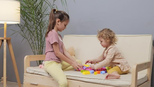 Happy kids cute sibling playing with colorful toys together while sitting on sofa little girls creating colorful figures with blocks enjoying funny playful time together